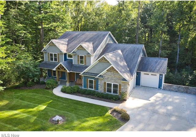 craftsman house featuring a front yard, roof with shingles, concrete driveway, stone siding, and a view of trees