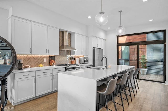 kitchen featuring wall chimney range hood, white cabinets, hanging light fixtures, an island with sink, and light wood-type flooring