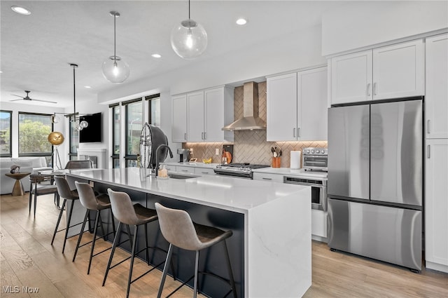 kitchen featuring wall chimney range hood, an island with sink, white cabinets, appliances with stainless steel finishes, and light hardwood / wood-style floors