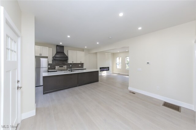 kitchen with light wood-type flooring, white cabinets, custom range hood, a center island with sink, and appliances with stainless steel finishes