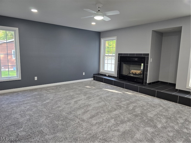 unfurnished living room with a tile fireplace, dark colored carpet, ceiling fan, and a wealth of natural light