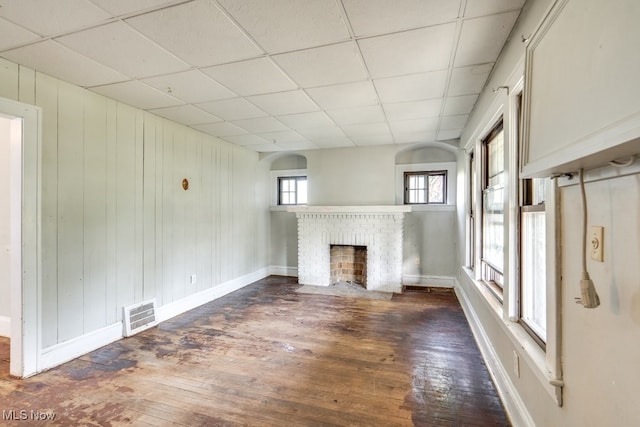 unfurnished living room featuring a paneled ceiling, wood walls, dark wood-type flooring, and a brick fireplace