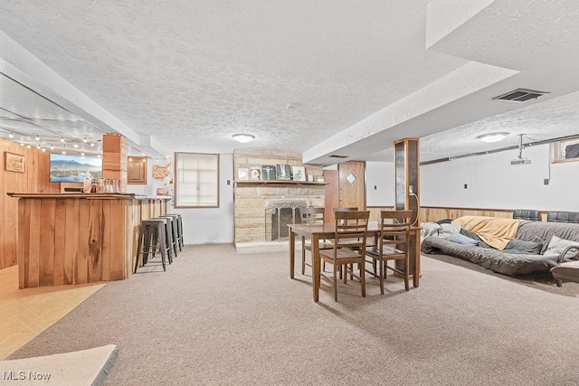 dining area with a textured ceiling, bar area, light colored carpet, wooden walls, and a stone fireplace