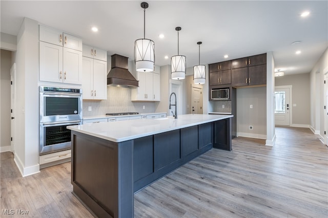 kitchen with a kitchen island with sink, light hardwood / wood-style floors, white cabinetry, custom exhaust hood, and appliances with stainless steel finishes