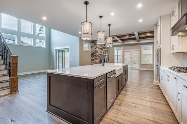 kitchen with light wood-type flooring, white cabinets, hanging light fixtures, a center island with sink, and beam ceiling