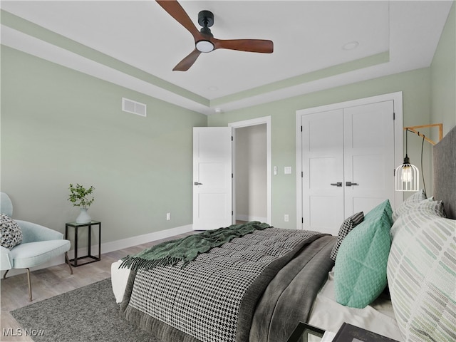 bedroom with ceiling fan, light wood-type flooring, a tray ceiling, and a closet