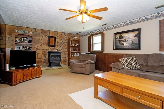 carpeted living room with a textured ceiling, wooden walls, ceiling fan, and a wood stove