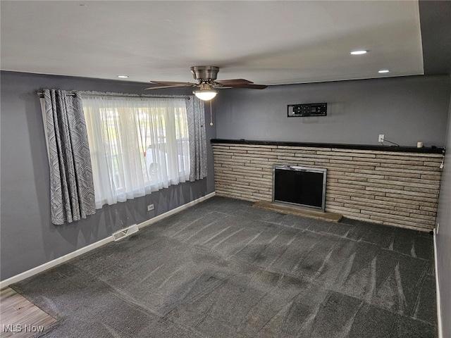 unfurnished living room with dark colored carpet, ceiling fan, and a stone fireplace