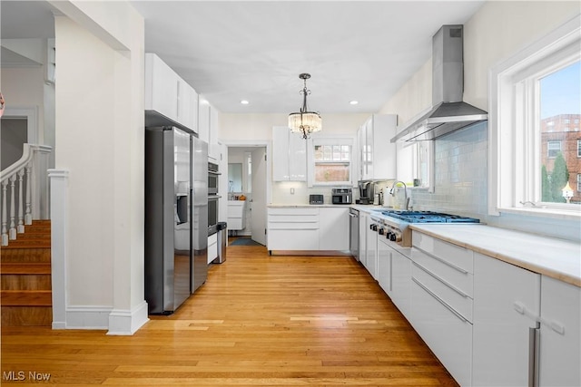 kitchen with wall chimney range hood, a wealth of natural light, appliances with stainless steel finishes, light wood-style floors, and white cabinets