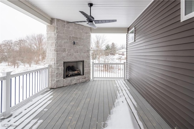 snow covered deck with ceiling fan and an outdoor stone fireplace