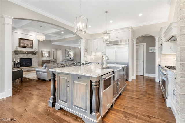 kitchen with hanging light fixtures, a kitchen island with sink, white cabinetry, high end stainless steel range oven, and ornate columns