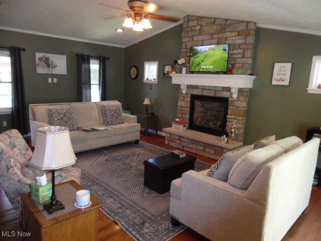 living room featuring wood-type flooring, vaulted ceiling, crown molding, a stone fireplace, and ceiling fan