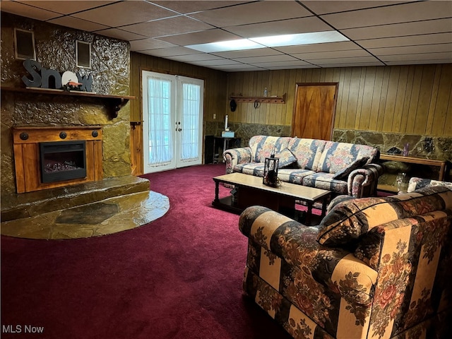 carpeted living room with a drop ceiling, wooden walls, a fireplace, and french doors