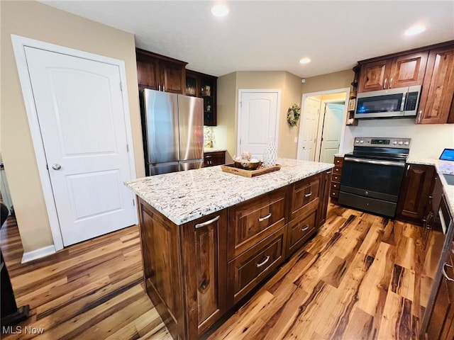 kitchen with appliances with stainless steel finishes, a center island, dark brown cabinetry, light stone counters, and light hardwood / wood-style floors