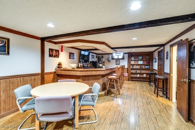 bar with light wood-type flooring, beamed ceiling, wooden walls, ornamental molding, and a textured ceiling