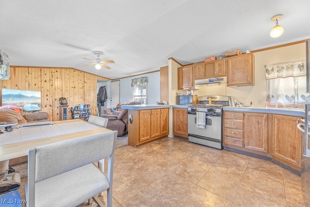 kitchen with kitchen peninsula, ceiling fan, wooden walls, stainless steel gas range oven, and ventilation hood