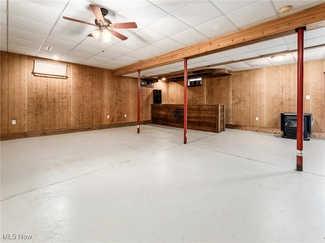 basement featuring wooden walls, a paneled ceiling, a wood stove, and ceiling fan