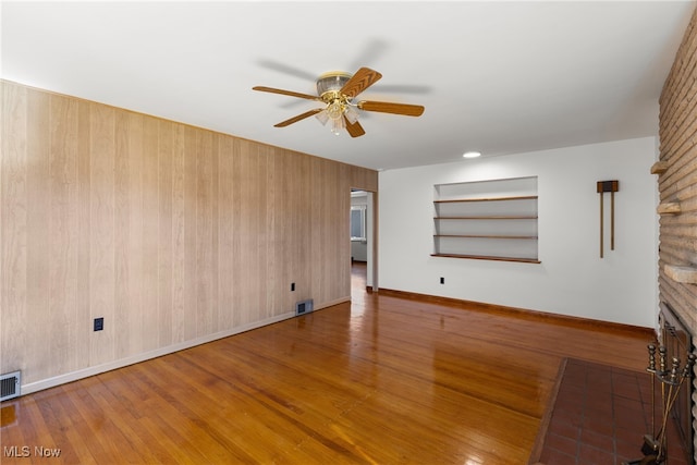 unfurnished living room featuring ceiling fan, wood-type flooring, a brick fireplace, built in shelves, and wooden walls