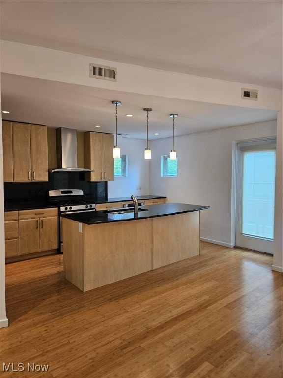 kitchen featuring an island with sink, gas range oven, hanging light fixtures, light hardwood / wood-style floors, and wall chimney exhaust hood