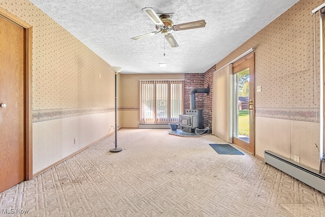 carpeted spare room featuring a wood stove, a baseboard heating unit, a textured ceiling, and a wealth of natural light