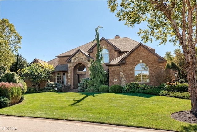 view of front facade featuring a front lawn, brick siding, roof with shingles, and a chimney