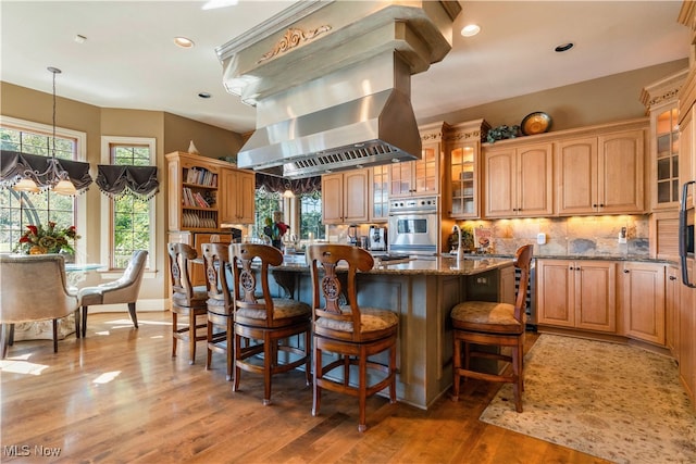 kitchen featuring island exhaust hood, a kitchen island with sink, pendant lighting, stone counters, and oven