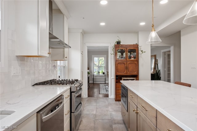 kitchen with wall chimney range hood, hanging light fixtures, white cabinets, appliances with stainless steel finishes, and light stone counters