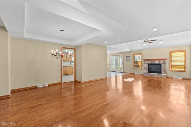 unfurnished living room with a tray ceiling, a fireplace, ceiling fan with notable chandelier, and light wood-type flooring