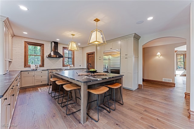 kitchen featuring hanging light fixtures, light hardwood / wood-style floors, high quality appliances, wall chimney exhaust hood, and a kitchen island with sink