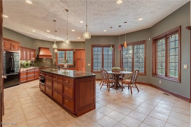 kitchen with tasteful backsplash, black fridge, custom range hood, decorative light fixtures, and an island with sink