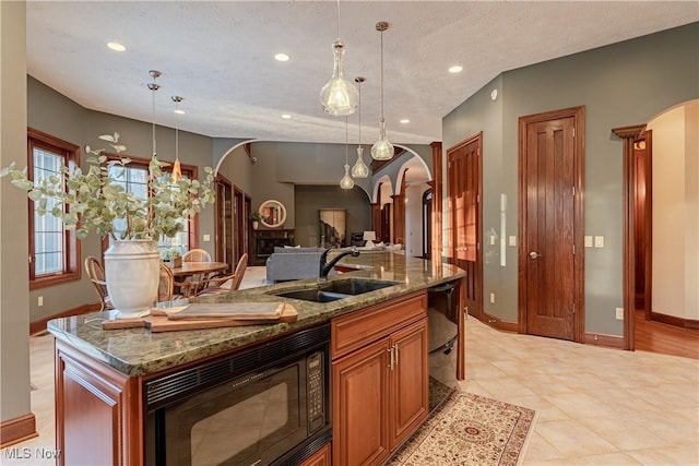 kitchen featuring black microwave, sink, stone countertops, decorative light fixtures, and an island with sink