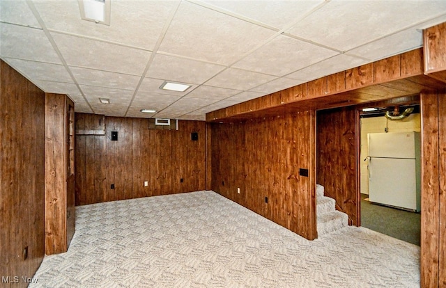 basement with a drop ceiling, wooden walls, light carpet, and white refrigerator