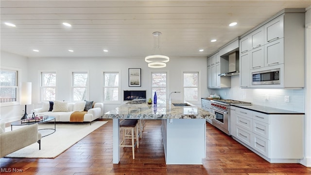 kitchen with appliances with stainless steel finishes, a large island with sink, dark hardwood / wood-style flooring, light stone countertops, and wall chimney exhaust hood