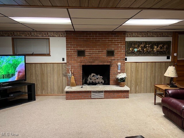 living room featuring wooden walls, a drop ceiling, carpet floors, and a brick fireplace