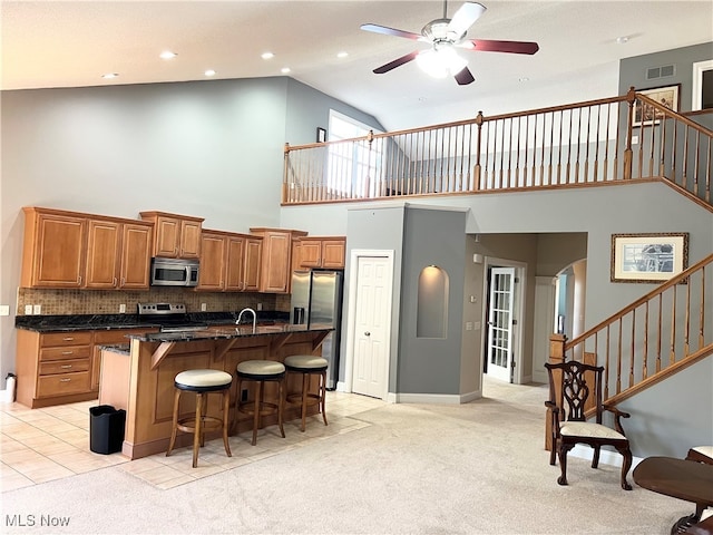 kitchen featuring light carpet, appliances with stainless steel finishes, ceiling fan, a center island with sink, and high vaulted ceiling