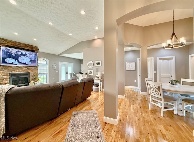 living room featuring a stone fireplace, a notable chandelier, light hardwood / wood-style floors, vaulted ceiling, and a textured ceiling