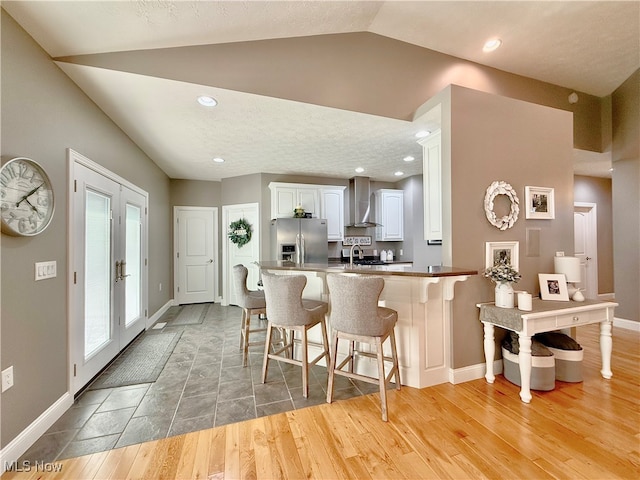 kitchen with kitchen peninsula, stainless steel fridge, wall chimney range hood, white cabinetry, and lofted ceiling