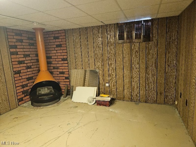 basement featuring brick wall, a paneled ceiling, a wood stove, and wood walls