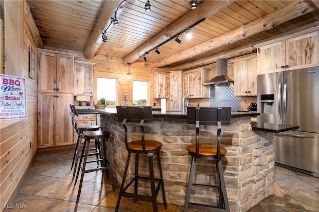 kitchen featuring a breakfast bar, wall chimney range hood, rail lighting, stainless steel fridge, and beamed ceiling