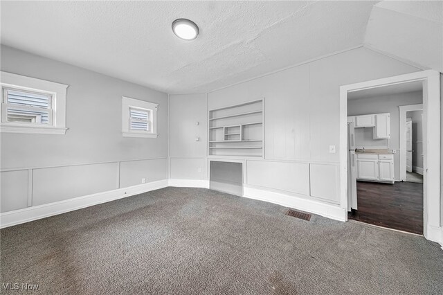 unfurnished living room with a decorative wall, a textured ceiling, built in features, dark carpet, and visible vents