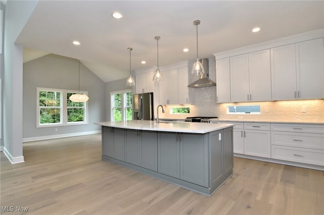 kitchen featuring lofted ceiling, white cabinets, wall chimney exhaust hood, stainless steel fridge, and an island with sink