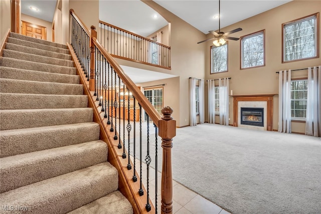 stairs featuring ceiling fan, a towering ceiling, a glass covered fireplace, tile patterned floors, and carpet flooring