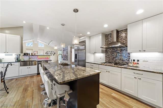 kitchen with sink, wall chimney exhaust hood, stainless steel appliances, an island with sink, and vaulted ceiling