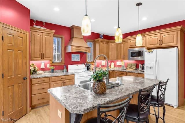 kitchen featuring hanging light fixtures, white appliances, a kitchen island with sink, custom exhaust hood, and light wood-type flooring