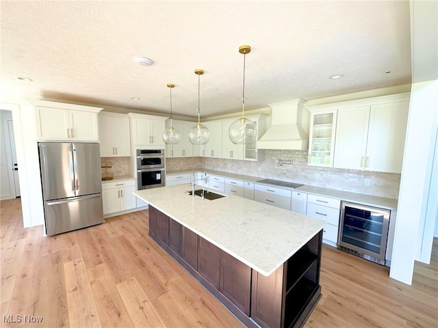 kitchen featuring a center island with sink, sink, custom range hood, stainless steel appliances, and beverage cooler