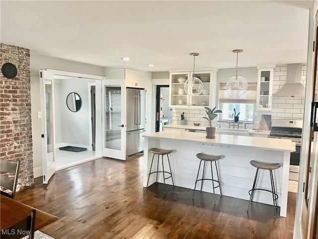 kitchen featuring high end refrigerator, white range oven, wall chimney range hood, white cabinetry, and hanging light fixtures