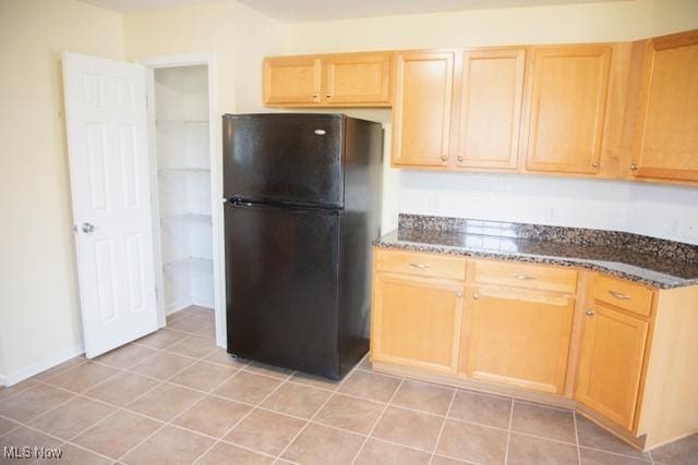 kitchen featuring black refrigerator, light tile patterned floors, light brown cabinetry, and dark stone countertops