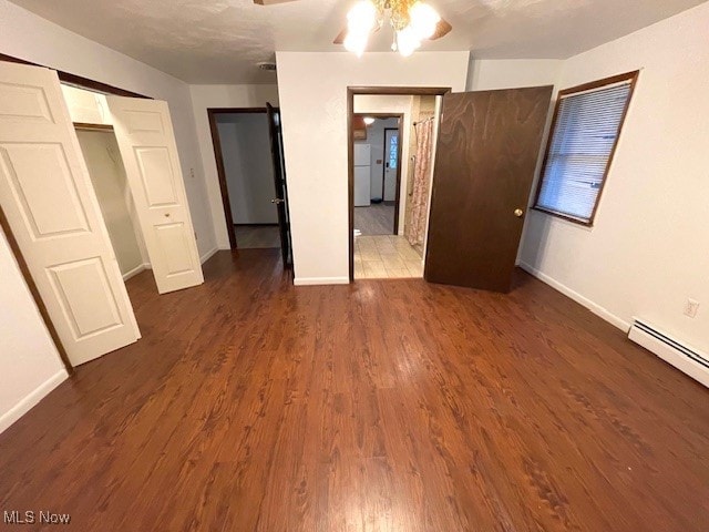 unfurnished bedroom with dark wood-type flooring, a baseboard heating unit, white refrigerator, ceiling fan, and a closet