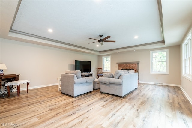 living room with light wood-type flooring, a tray ceiling, a brick fireplace, and ceiling fan