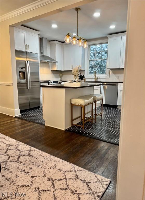 kitchen featuring white cabinetry, decorative light fixtures, appliances with stainless steel finishes, wall chimney range hood, and crown molding